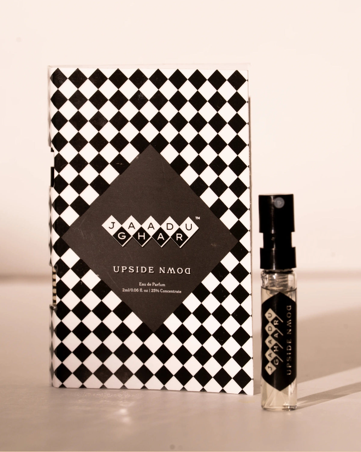 A small clear perfume spray bottle with a black cap stands beside its black-and-white checkered box. The front label reads 'JAADU GHARU' and 'UPSIDE DOWN'.