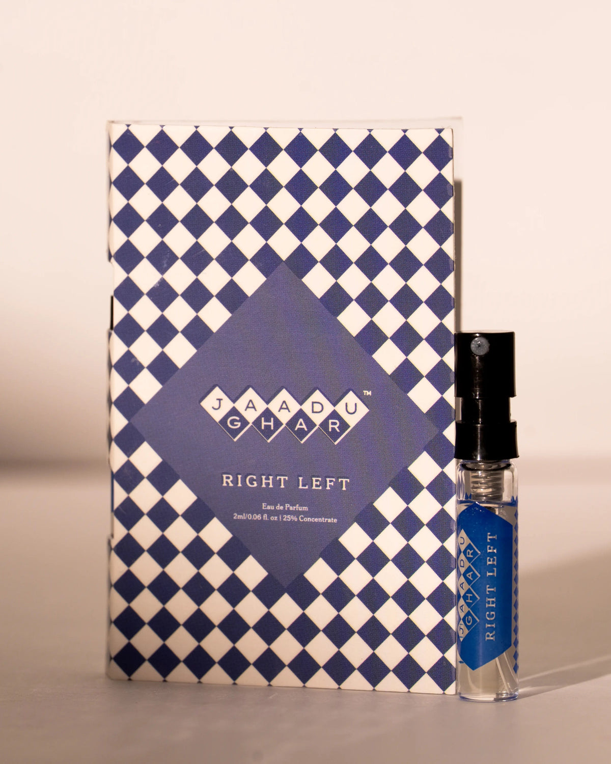 A small clear glass perfume sample vial with a black spray top stands beside its patterned retail box. The box and label use a blue-and-white harlequin diamond pattern, with the brand name Jaadu Ghar and the scent name Right Left visible.
